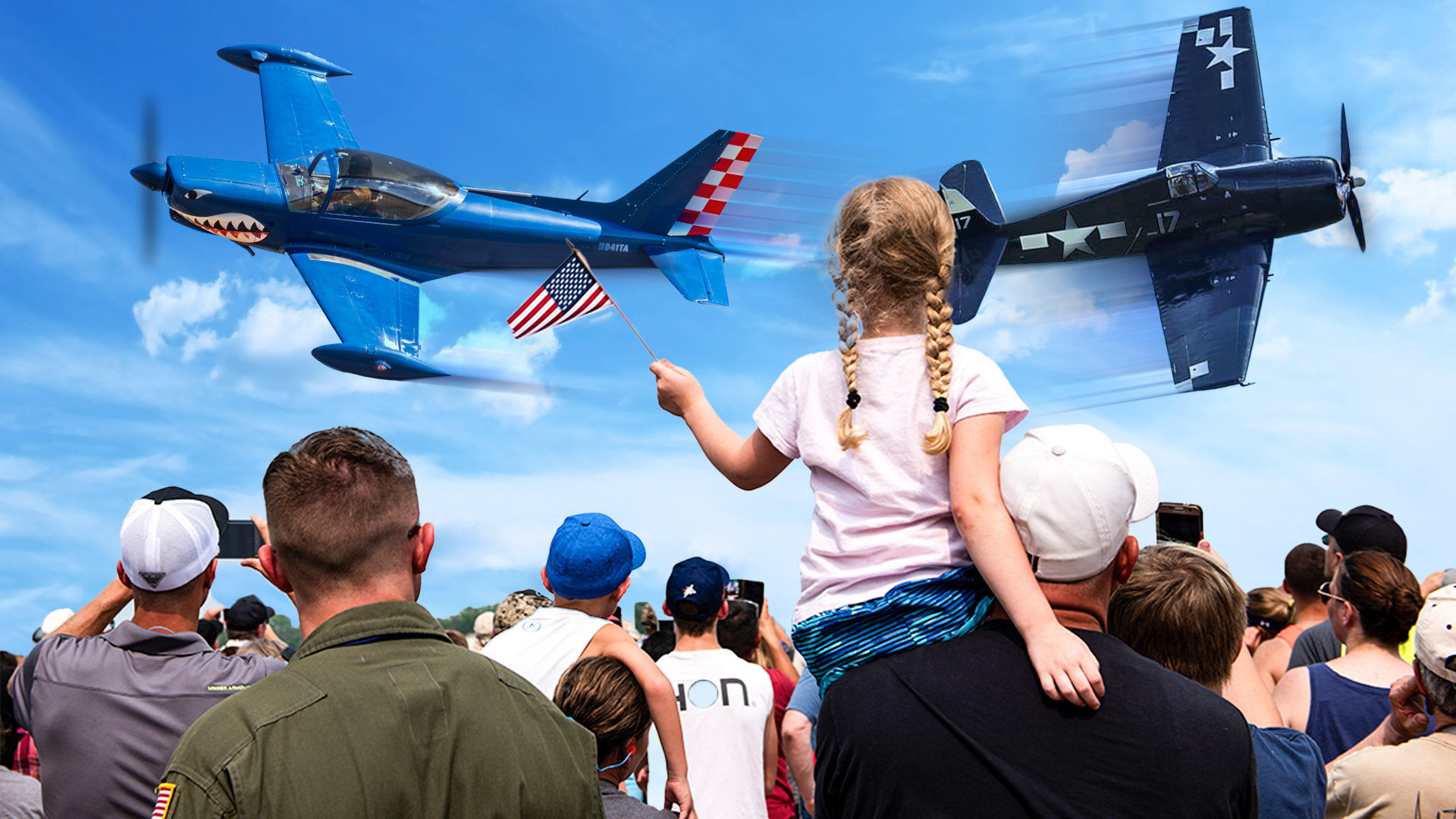 Young girl sitting on an adult’s shoulders waves a small American flag as two vintage military aircraft fly overhead during the 2026 Bremerton Air Show, with a large crowd watching and photographing the flyby.