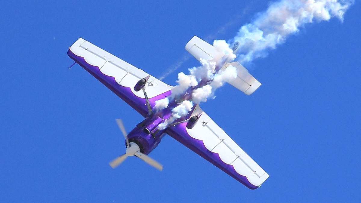 Aerobatic pilot Renny Price performing a maneuver in a purple and white Sukhoi SU-29 aircraft with smoke trailing from the wings during an air show performance against a clear blue sky.