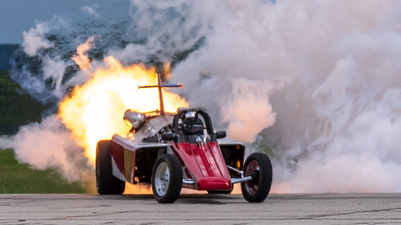 Smoke-n-Thunder Jet Car driven by Bill Braack performs on a runway, accelerating with a large burst of flame shooting from the rear jet engine and thick white smoke billowing behind. The red, open-cockpit jet-powered dragster has exposed front wheels and a low, narrow body, with the driver visible wearing a helmet inside the cockpit. The vehicle is angled slightly toward the viewer as it speeds across the tarmac, with the intense fire plume illuminating the back of the car and contrasting against the dense smoke cloud.