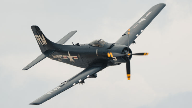 Douglas AD-4N Skyraider warbird operated by the Soaring by the Sea Foundation flying with landing gear retracted, painted in dark navy blue with U.S. Navy markings, visible propeller motion, and tail code RM on the vertical stabilizer.