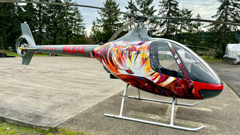 Cabri G2 helicopter by Pureflight Aviation parked on a concrete pad, featuring a colorful custom paint design in red, orange, and white tones, with tail number N484E visible, surrounded by trees and open sky in the background