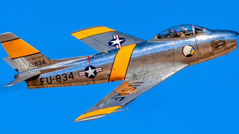 Polished silver F-86F Sabre jet owned by Planes of Fame flying against a clear blue sky, featuring yellow wing and fuselage bands, U.S. Air Force markings, and tail number FU-834 visible on the fuselage.
