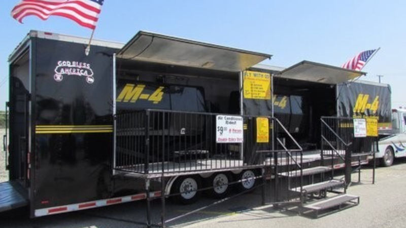 Black trailer setup housing the M-4 Flight Simulator with open side panels, stairs, and railings for entry, displaying signage and pricing information, with American flags mounted on top at an outdoor event setting
