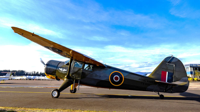 1944 Stinson AT-19 aircraft from the CAF Rainier Squadron parked on an airport ramp, side view showing dark olive paint, Royal Navy roundel, and high-wing design under a blue sky.