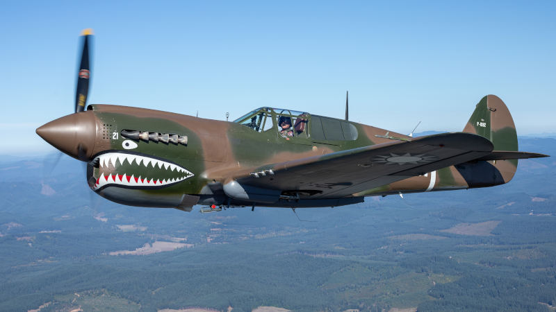 Curtiss P-40 Kittyhawk warbird operated by the Soaring by the Sea Foundation flying over a landscape, painted in green and brown camouflage with shark mouth nose art, U.S. military markings, and a pilot visible in the cockpit.