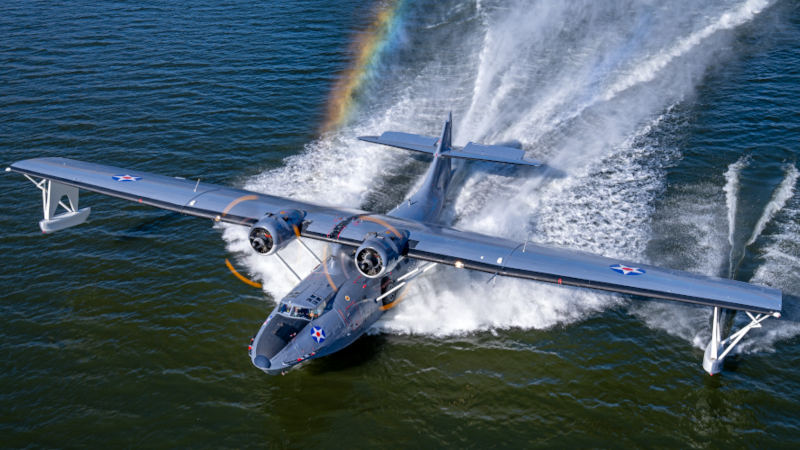 PBY-5A Catalina amphibious aircraft operated by the Soaring by the Sea Foundation lifting off from the water, creating large spray trails behind its hull and wingtip floats, with U.S. military markings visible and a rainbow forming in the mist above the wake.