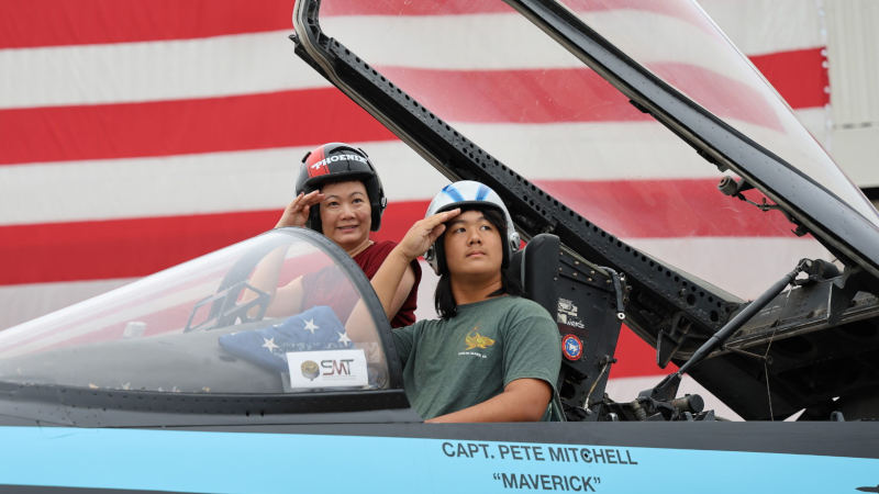 Two participants wearing flight helmets sit in a mock F/A-18 cockpit labeled “Capt Pete Mitchell ‘Maverick’,” saluting while seated under an open canopy, with a large American flag backdrop during the Top Gun cockpit experience at the 2026 Bremerton Air Show.