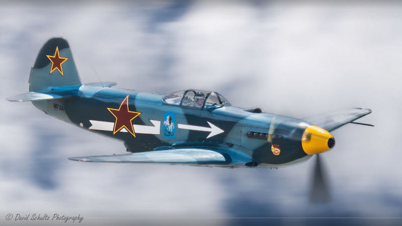 Yak-3 WWII-era fighter aircraft owned by Bill Sheppard flying at speed with blurred propeller, painted in teal with red star insignia and yellow nose cone against a soft, motion-blurred background.