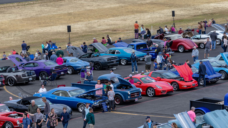 Classic cars and modern muscle cars line the tarmac during the Bremerton Airshow classic car show in Washington state as visitors walk between rows of vehicles with open hoods on display.