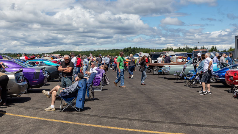 Spectators gather among rows of classic cars and restored vehicles during the Bremerton Airshow classic car show in Washington state under a partly cloudy sky.