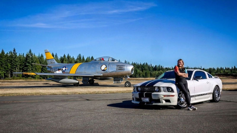 Woman leaning against a white Ford Mustang during a planeside drive-up photo session with a restored F-86 Sabre jet parked on the tarmac behind her.