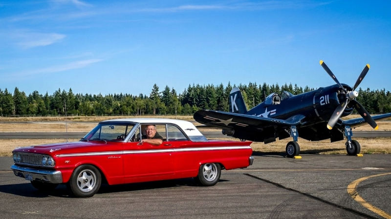 Driver posing in a red vintage Ford Fairlane during a planeside drive-up photo session beside a restored F4U Corsair warbird on the airport ramp.