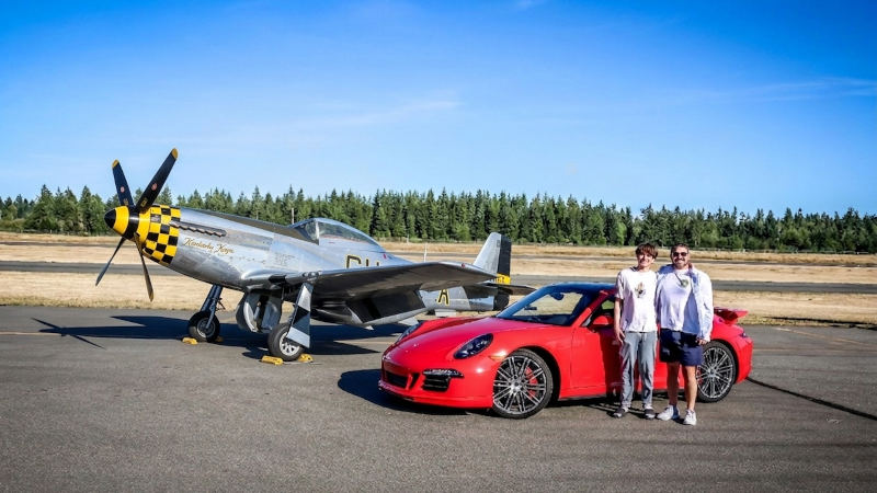 Two guests standing beside a red Porsche during a planeside drive-up photo session with a restored P-51D Mustang parked on the airport ramp.