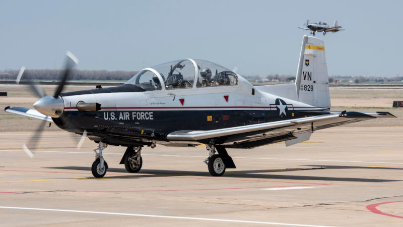 U.S. Air Force T-6 Texan II training aircraft from Vance Air Force Base taxiing on the airport ramp with its propeller spinning. The two-seat turboprop is shown in side profile wearing the U.S. Air Force's white, dark blue, and silver training aircraft livery, with the tandem cockpit canopy closed and pilots visible inside. Another aircraft is visible in the background above the tail.