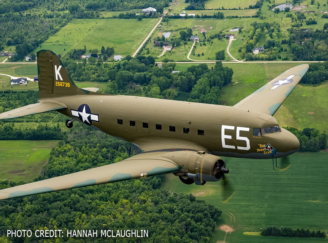 A vintage military transport aircraft flies over green farmland and wooded areas, with its olive drab paint scheme, U.S. Army Air Forces markings, and “The Yacht Club” nose art visible.