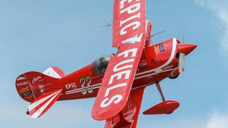 A red aerobatic biplane with “EPIC FUELS” branding flies against a blue sky, with its propeller spinning and the number 33 visible on the fuselage.