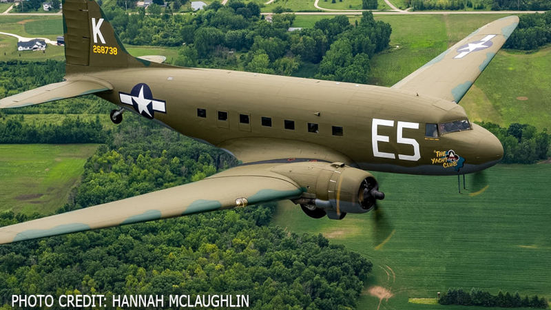 A vintage military transport aircraft flies over green farmland and wooded areas, with its olive drab paint scheme, U.S. Army Air Forces markings, and “The Yacht Club” nose art visible.